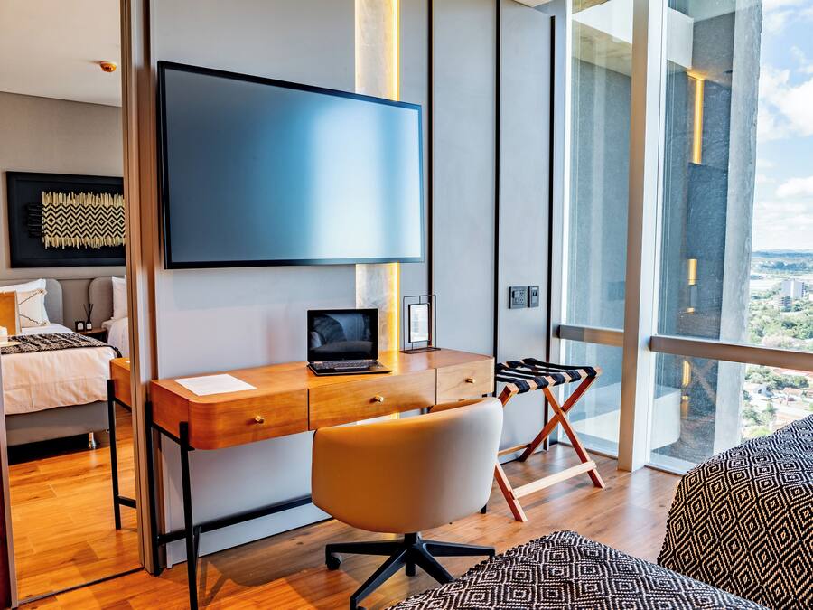 A modern bedroom with a wooden desk, a laptop, a TV, and a large window offering a city view.