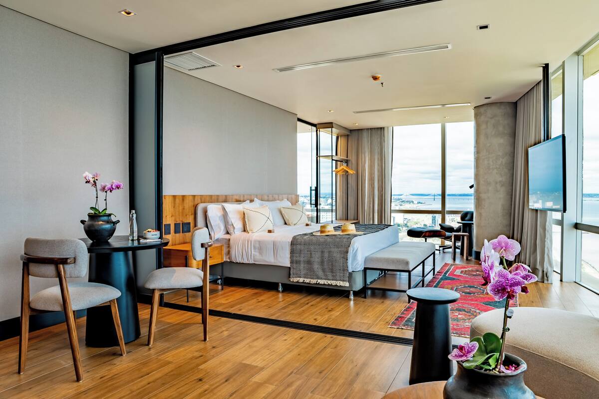 A modern hotel room with a large bed, a seating area with chairs, a small table, and a view of the city through large windows.