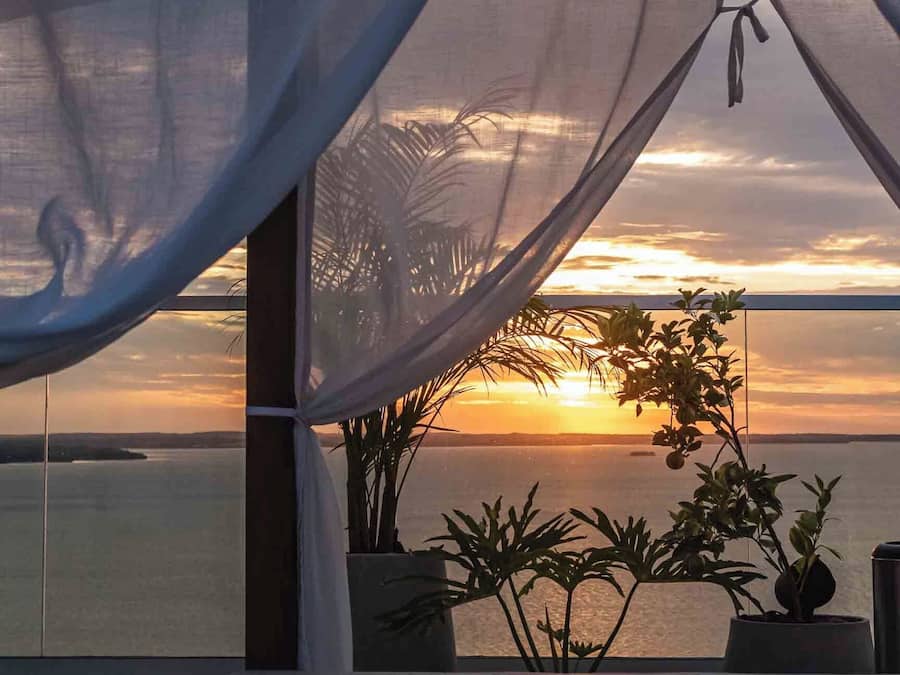 A room with a view of the sunset over the water, a balcony with potted plants, and a sheer curtain.