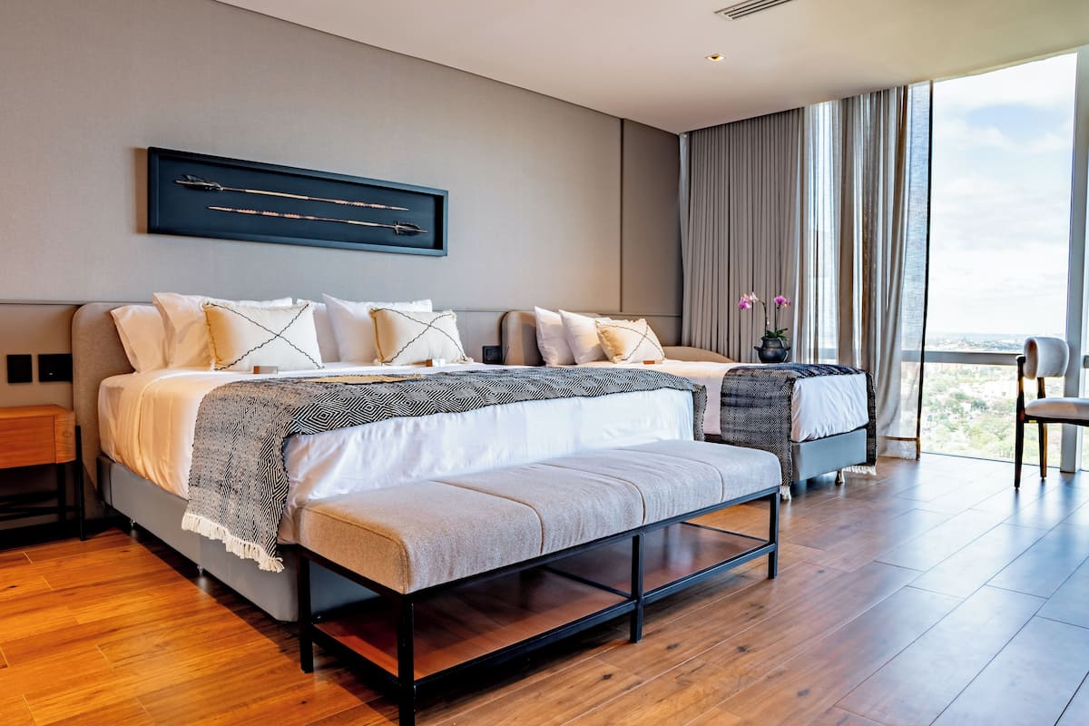 A modern bedroom with a large bed, a smaller bed, and a bench. There is a large window with curtains, a wall-mounted artwork, and a view of the city.