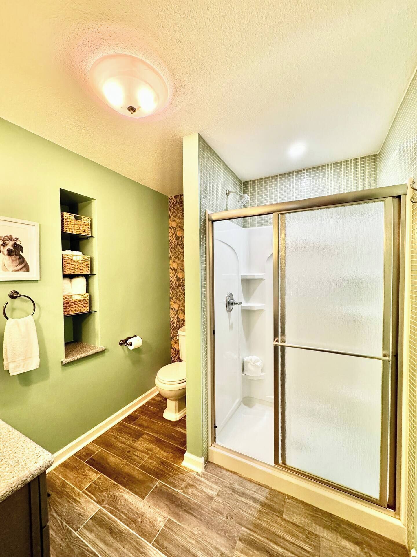 Un baño con pared verde, suelo de madera, inodoro, ducha envuelta en vidrio y un estante con cestas.