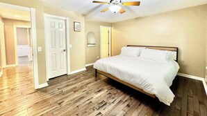 Un dormitorio con cama, suelo de madera, ventilador de techo y una puerta que da a otra habitación.