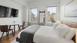 A modern hotel room with a bed, a desk, and a chair. There are two windows with a city view.