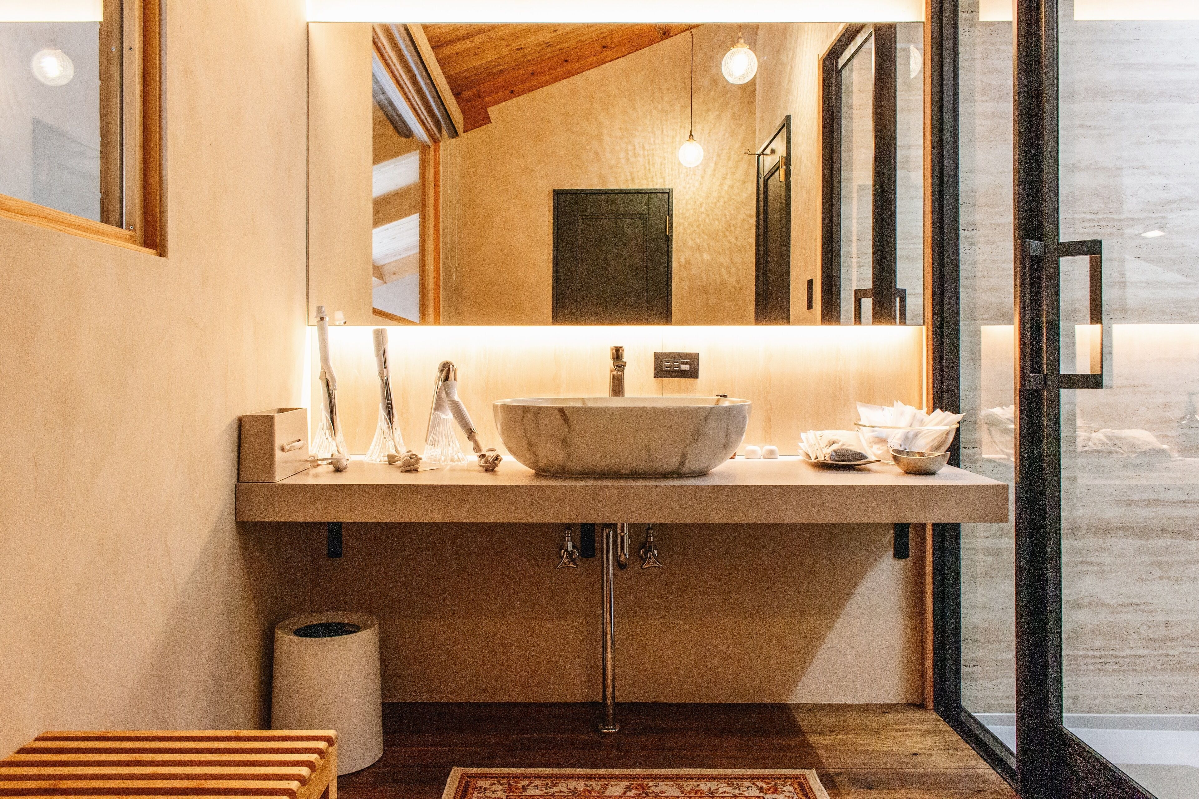 Un baño moderno con un lavamanos grande de mármol, un banco de madera y vistas a través de una puerta de cristal.