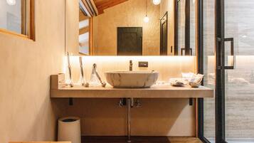 Un baño moderno con un lavamanos grande de mármol, un banco de madera y vistas a través de una puerta de cristal.