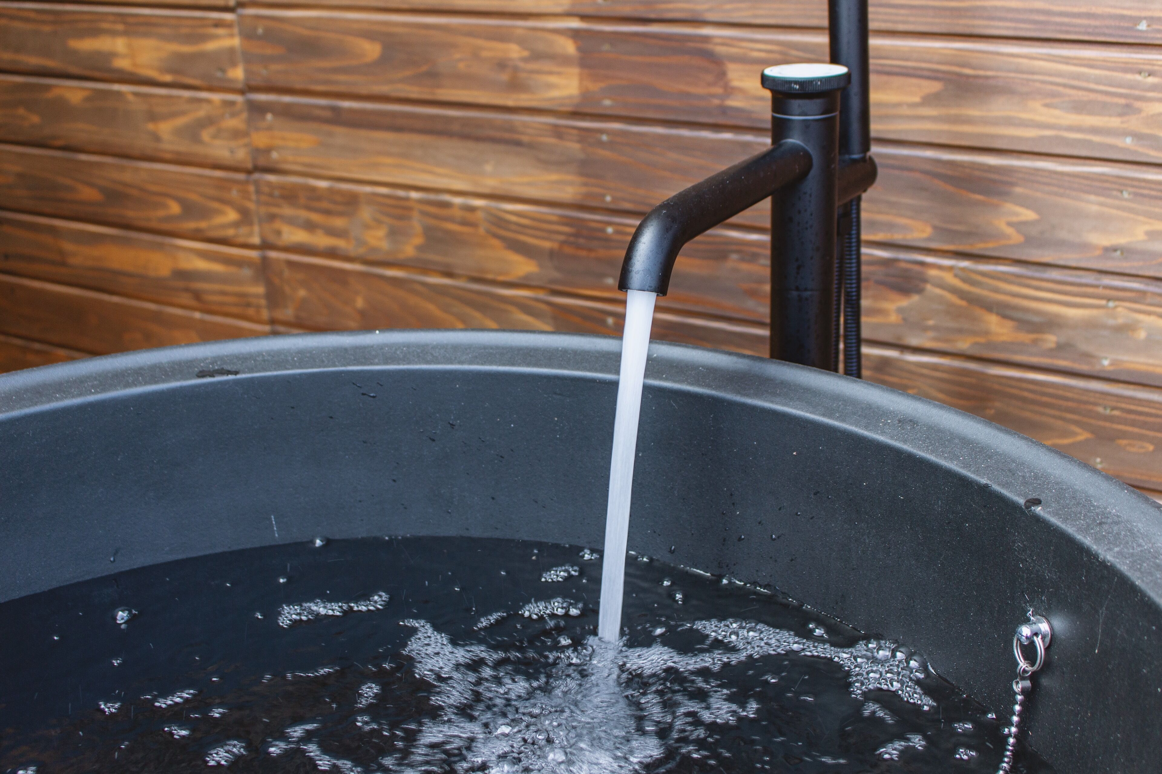 Eine große schwarze Badewanne mit Wasser, das aus einem Wasserhahn fließt, vor einer Holzwand.