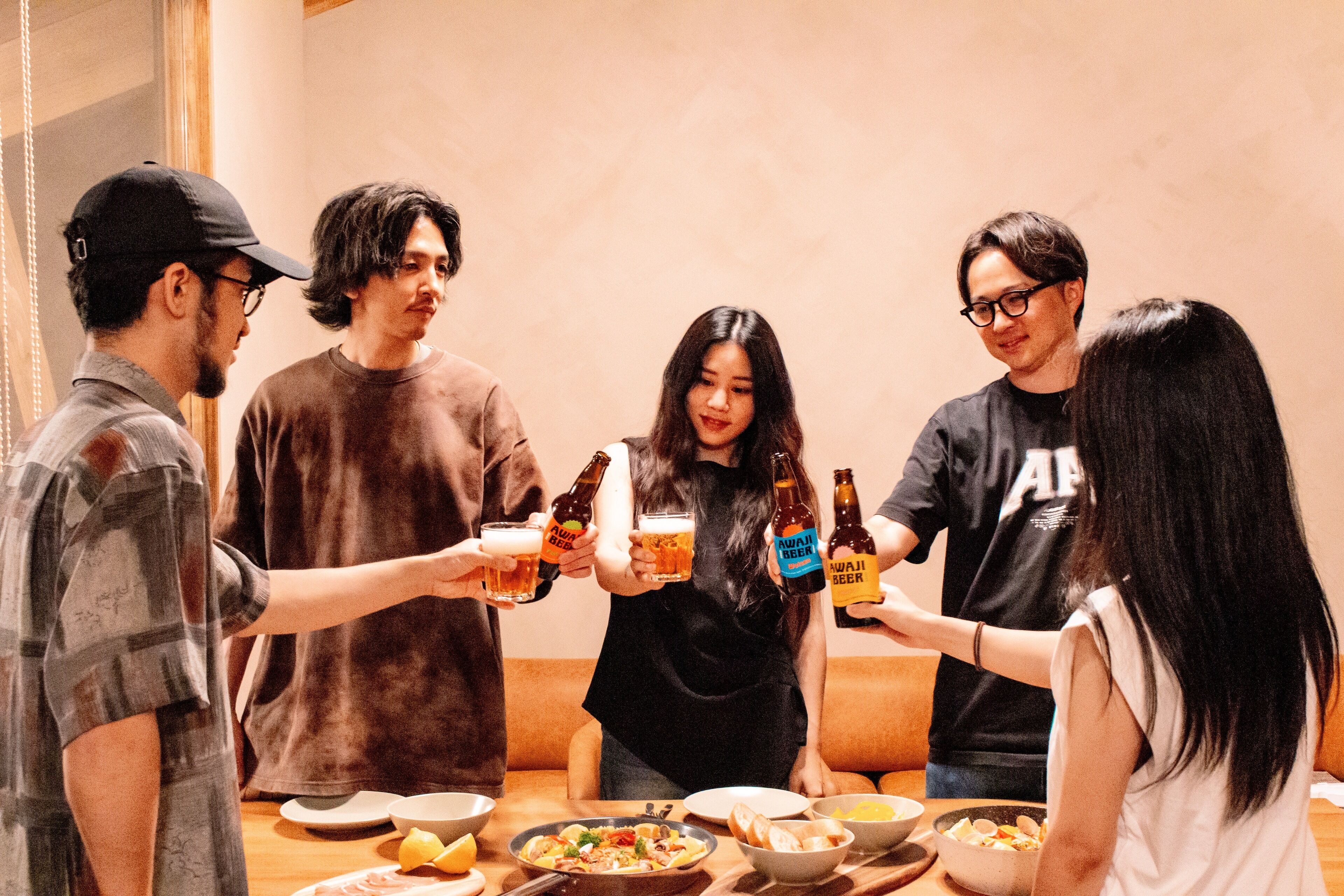 Un groupe de personnes trinquant avec des bouteilles de bière autour d’une table où se trouvent de la nourriture et des bols.