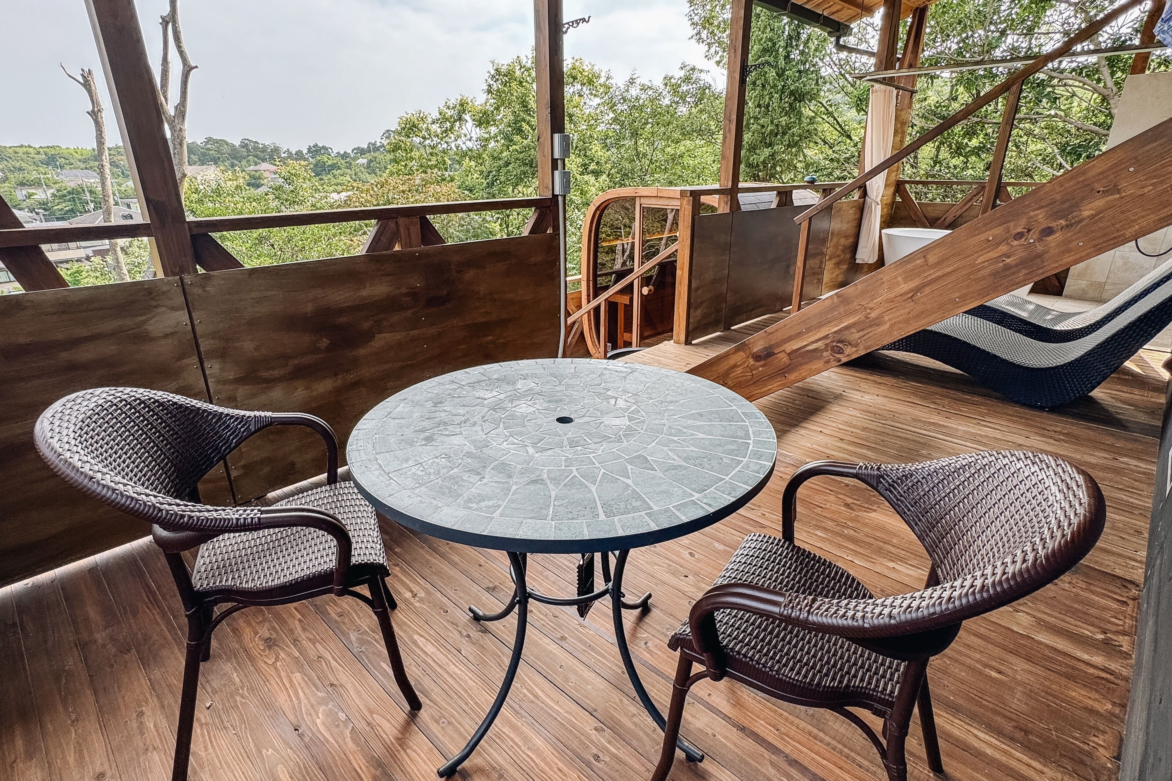 Una terraza de madera con una mesa redonda y sillas de mimbre, con vistas a un paisaje verde exuberante.