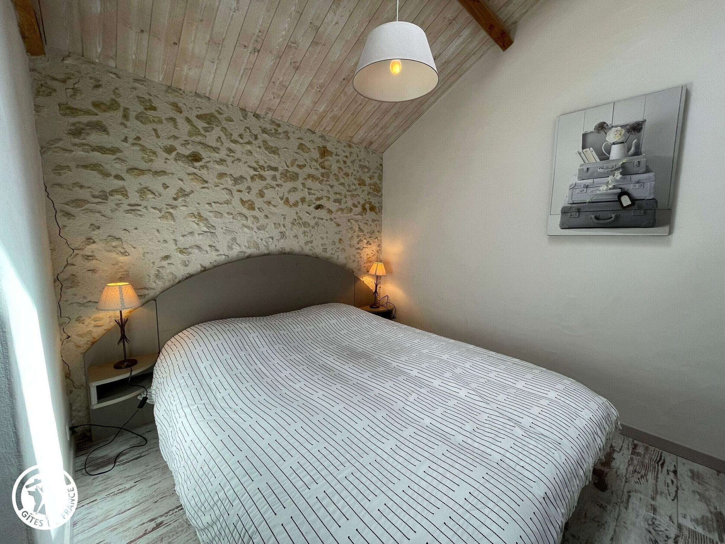 A bedroom with a stone wall, a bed with a patterned bedspread, two bedside lamps, and a wall-mounted picture.