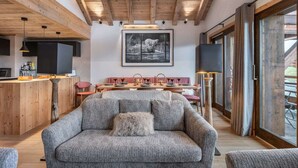 A cozy living room with a grey sofa, wooden ceiling beams, a dining area with pink chairs, and a large window.