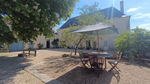 Un cortile con tavolo e sedie all'ombra di un ombrellone, un edificio con un camino e un albero.