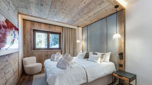 A modern bedroom with a wooden ceiling, a large bed with white bedding, a bedside table, and a window with a view of trees. - Exceptional apartment, ski-in, sauna, parking included (Méribel)