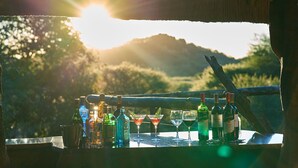 A table with various bottles and glasses, a metal pitcher, and a fork, set against a scenic outdoor view with a mountain in the background. - Sable Ranch Thaba Khaya (Brits)