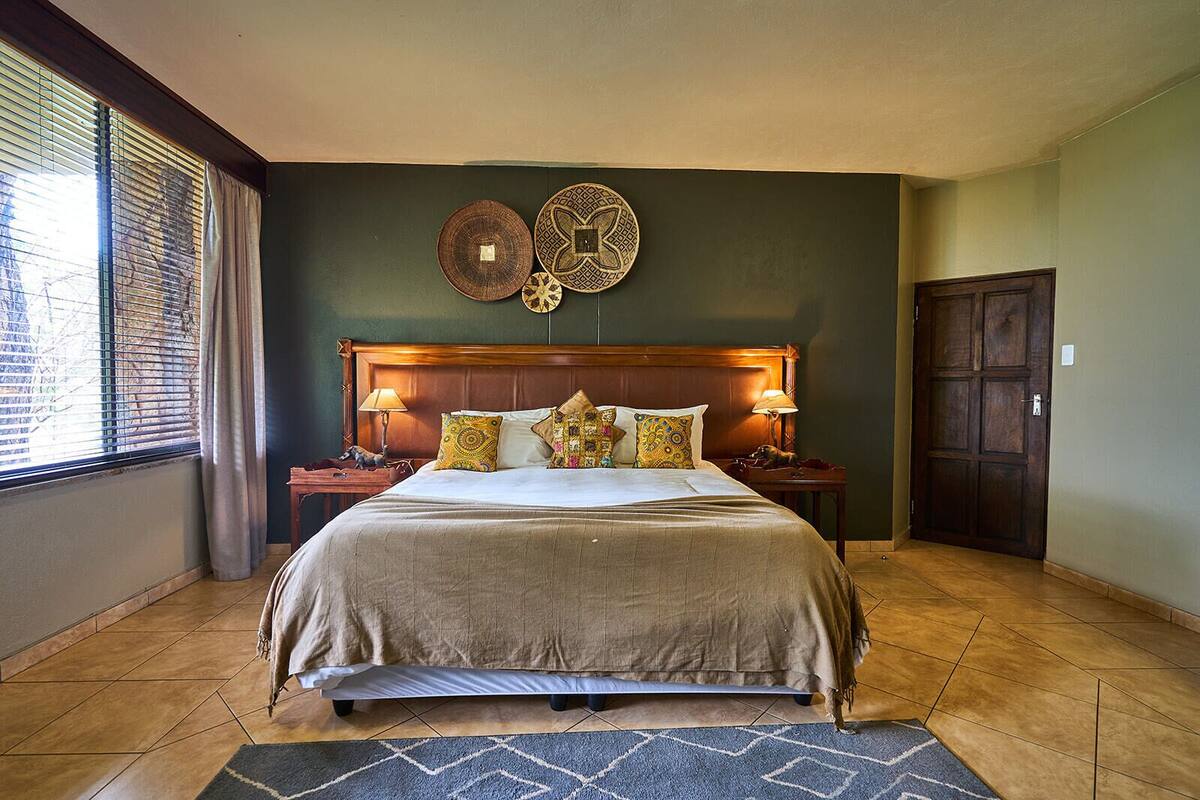 A bedroom with a large bed, two bedside tables, a wooden headboard, a window with blinds, and a door.