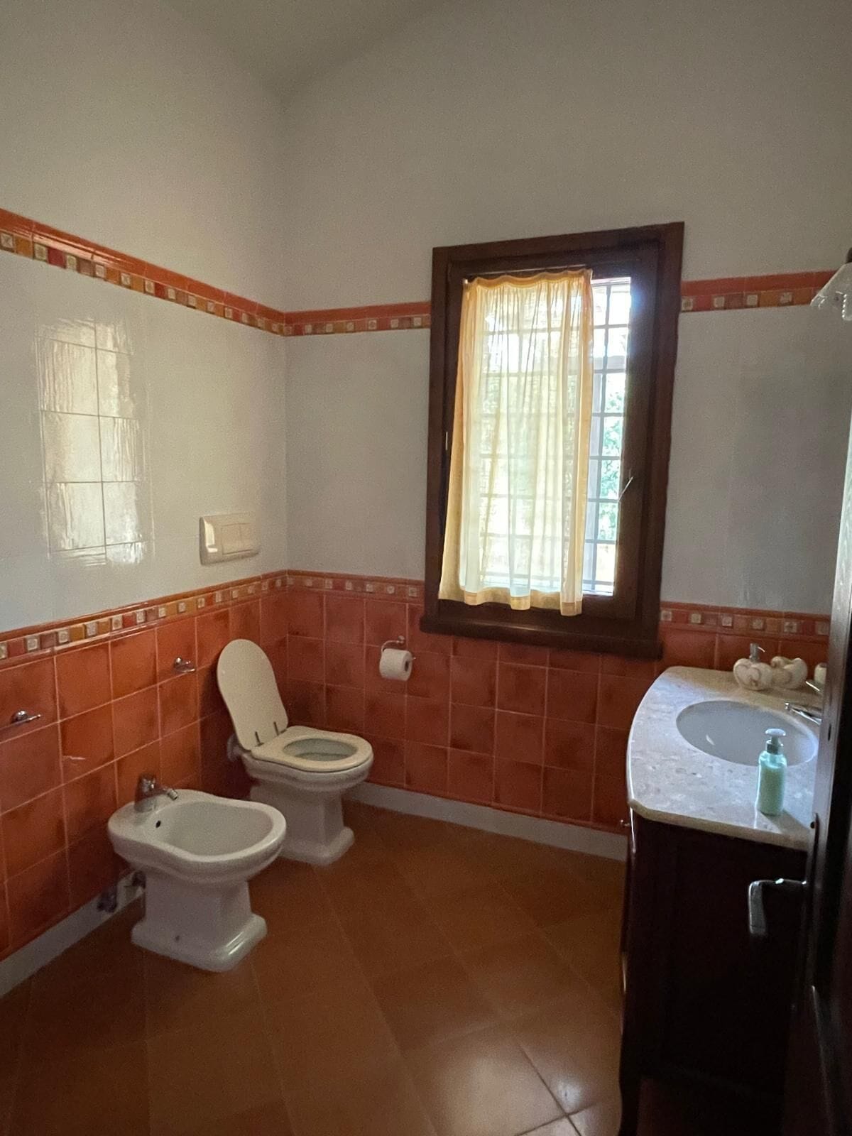 A bathroom with two toilets, a bidet, a sink, and a window with curtains.