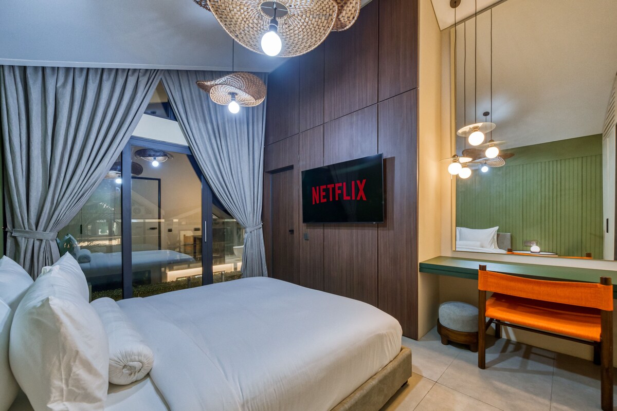 Une chambre d’hôtel moderne équipée d’un lit, d’un bureau, d’un téléviseur diffusant du contenu « NETFLIX » et d’une grande fenêtre avec des rideaux.