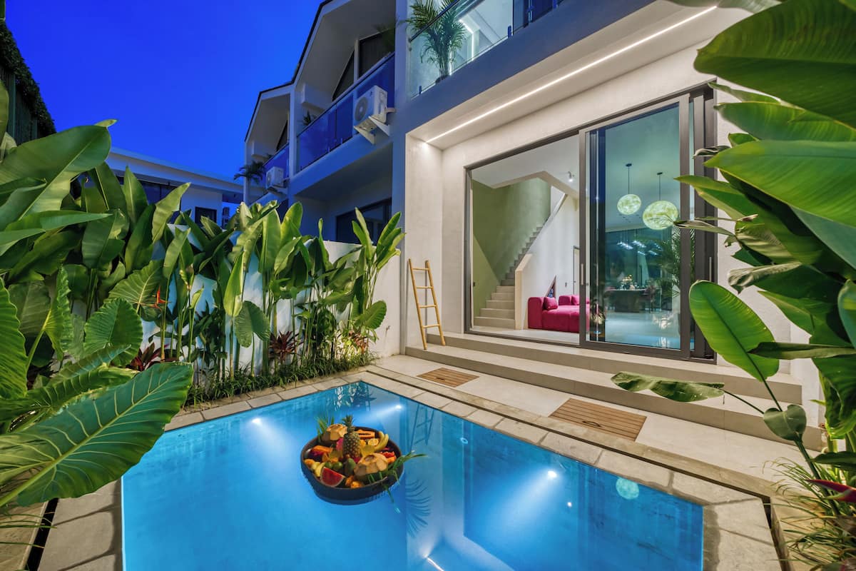 A modern pool area with a fruit bowl, surrounded by tropical plants and a glass door leading to a bright interior.
