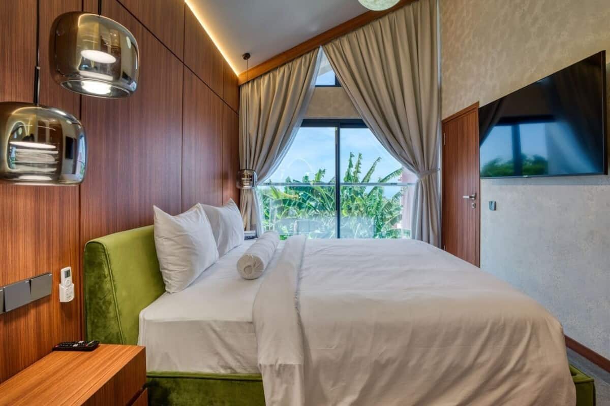 A hotel room with a large bed, wooden panel walls, a green headboard, and a view of greenery outside.