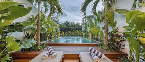 A poolside area with lounge chairs and a wooden deck leading to a pool, surrounded by tropical plants.