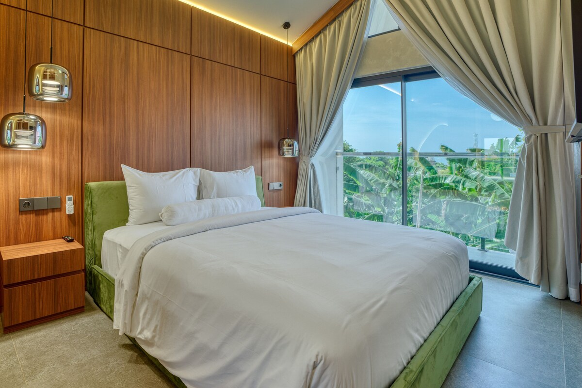 A modern bedroom with a large bed, wooden panel walls, a nightstand, and a view of greenery through a large window.