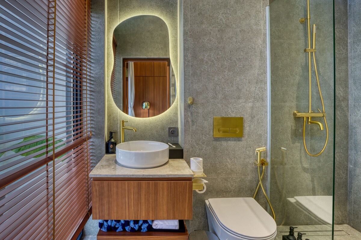 A modern bathroom with a round sink, a toilet, a shower with a golden showerhead, and a mirror with a wooden frame.