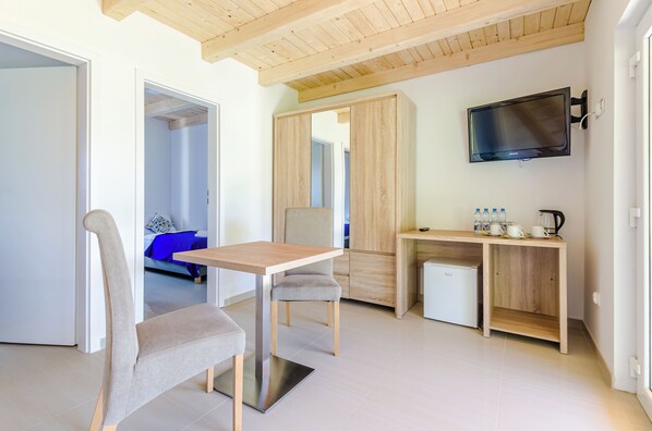A modern, well-lit room with a wooden ceiling, a TV mounted on the wall, a small kitchenette, and a seating area with a table and chairs.