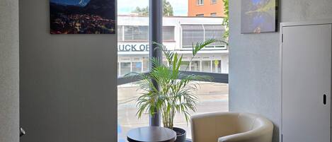 A hotel lobby with a large window, a potted plant, a round table, a sofa, and a wall-mounted picture.