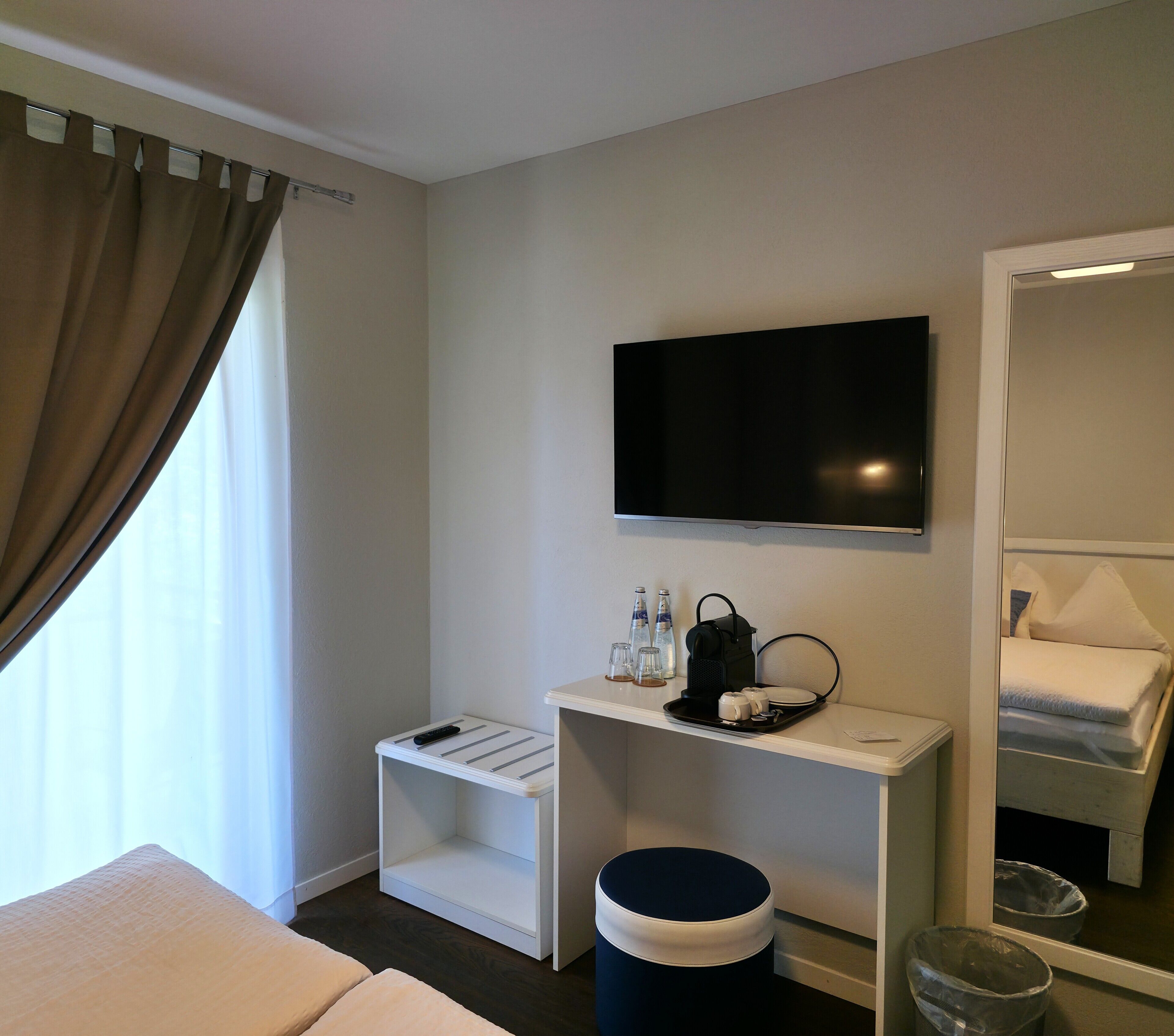 Ein Hotelzimmer mit Bett, einem an der Wand befestigten Fernseher, einem kleinen Tisch mit Tablett, einem Mülleimer und einem Fenster mit Vorhängen.