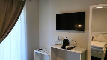 Ein Hotelzimmer mit Bett, einem an der Wand befestigten Fernseher, einem kleinen Tisch mit Tablett, einem Mülleimer und einem Fenster mit Vorhängen.