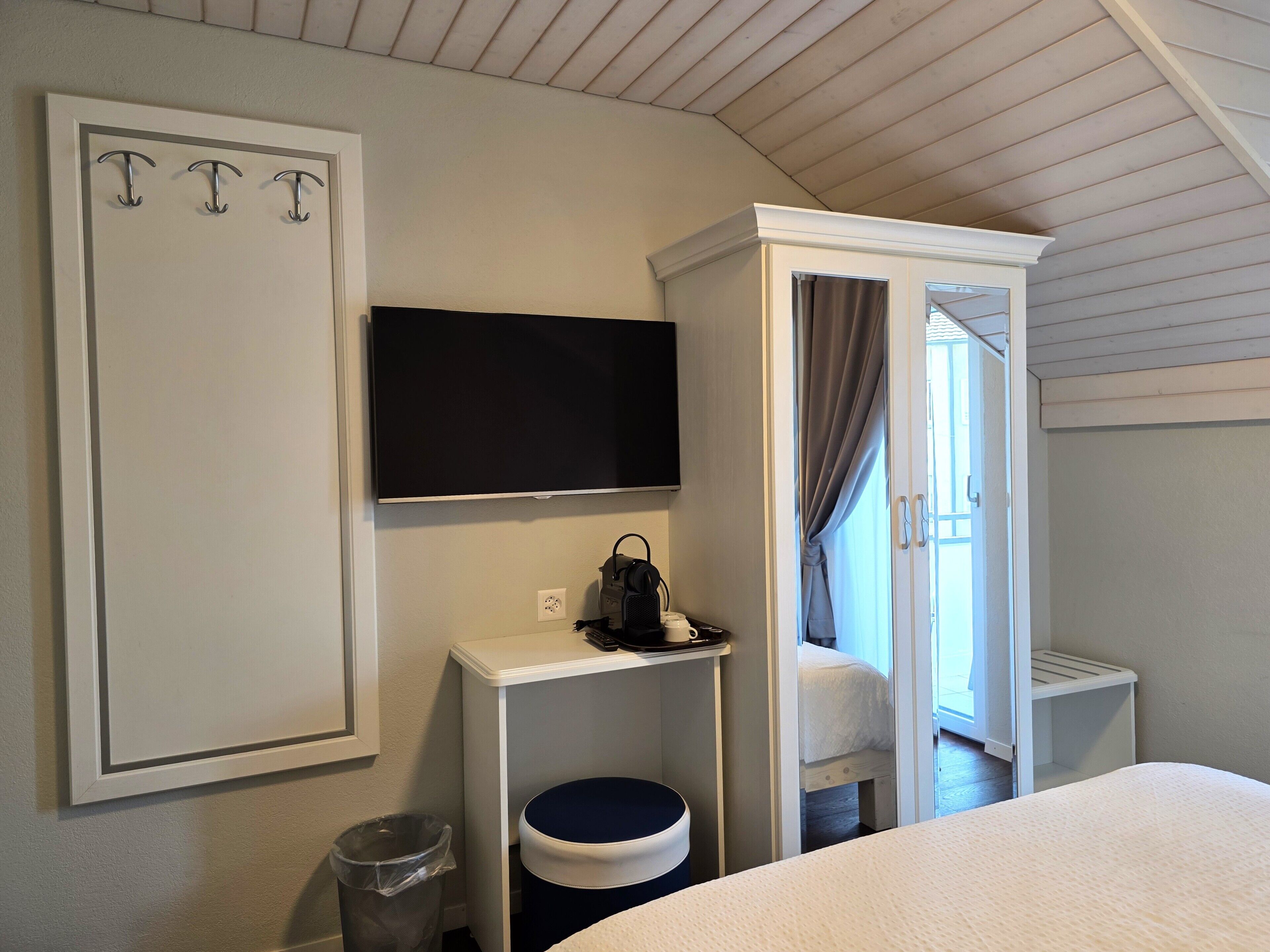 A hotel room with a bed, a TV mounted on the wall, a small table with a trash bin, and a wardrobe.