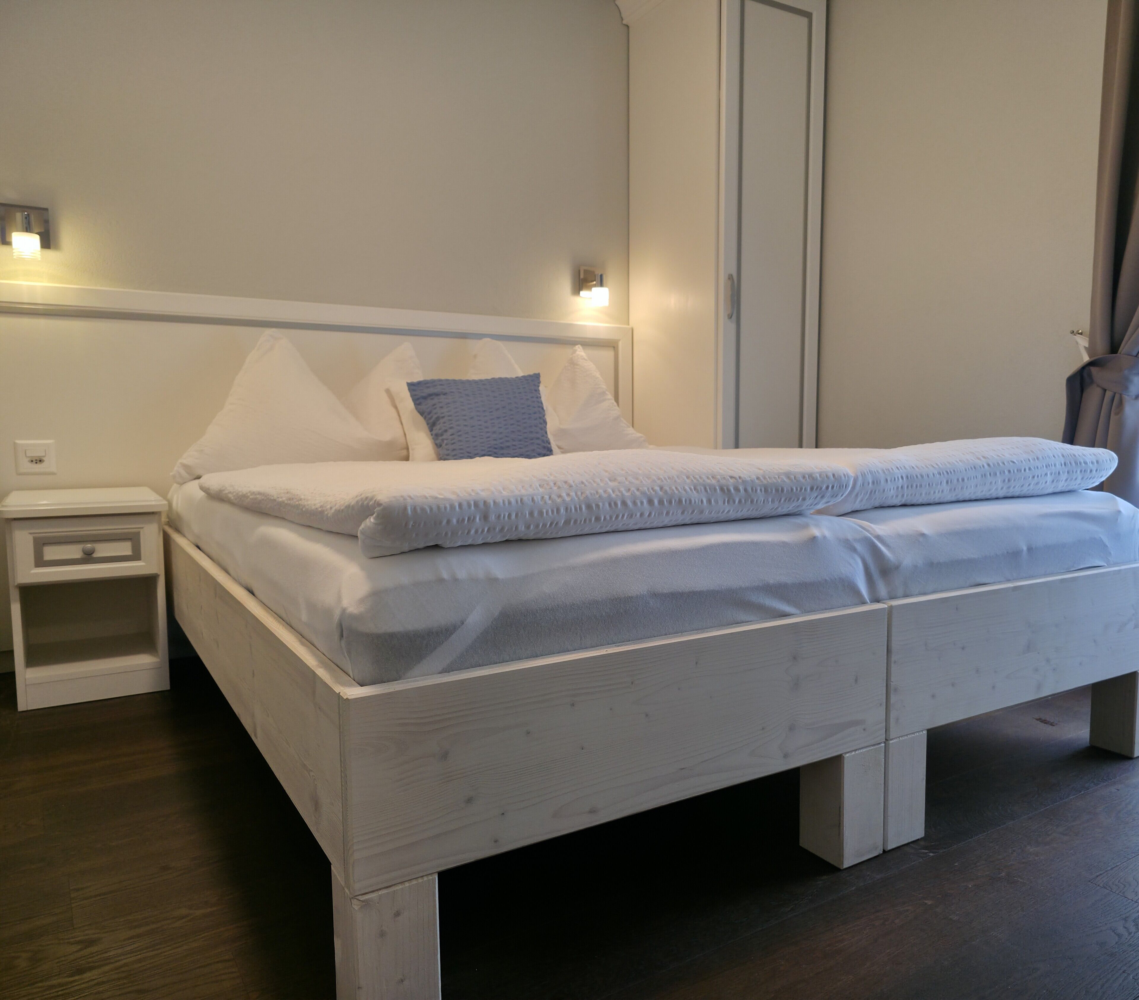 Una cama blanca con cabecera, una mesita de noche y una lámpara de pared.