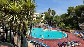 En pool omgiven av palmer och solstolar, med en byggnad i bakgrunden.