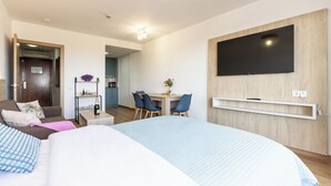Ett modernt hotellrum med en stor säng, ett skrivbord med stolar, en tv som är monterad på väggen och en dörr som leder till ett litet kök.