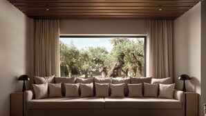 Un salon moderne avec un canapé beige, deux lampadaires noirs et une grande fenêtre donnant sur un paysage d’oliviers.