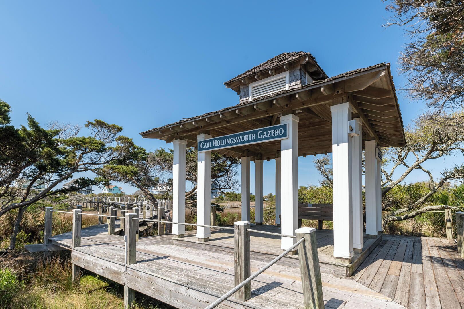 Een houten prieel met een bord waarop staat 'Carl Hollingsworth Gazebo', gelegen in een natuurlijke omgeving met bomen en een heldere lucht.