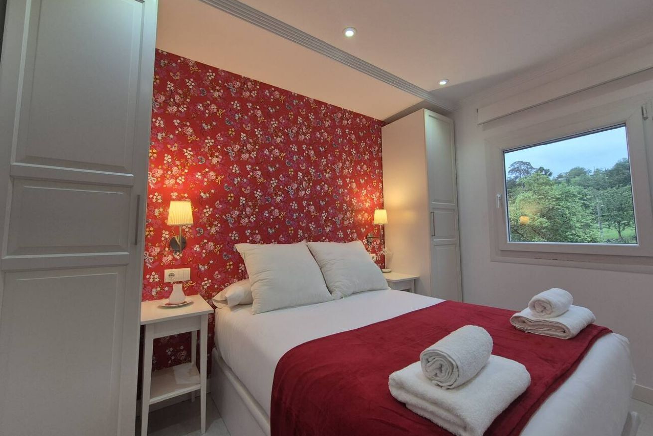 A bedroom with a floral wallpaper, a red bedspread, two bedside lamps, a window with a view of greenery, and neatly folded towels on the bed.