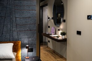 A modern hotel room with a large bed, a nightstand, a desk with a lamp, a mirror, and a small potted plant.