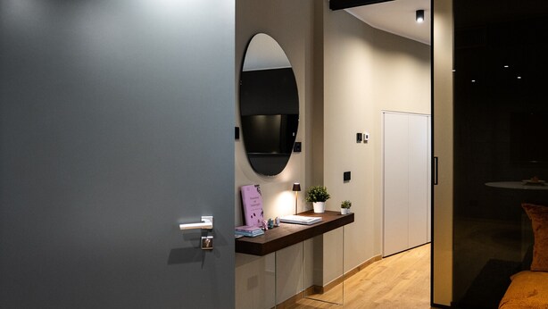 Ein modernes Hotelzimmer mit Holzboden, einem Schreibtisch mit Lampe, einem Blumentopf und einem großen Spiegel an der Wand.