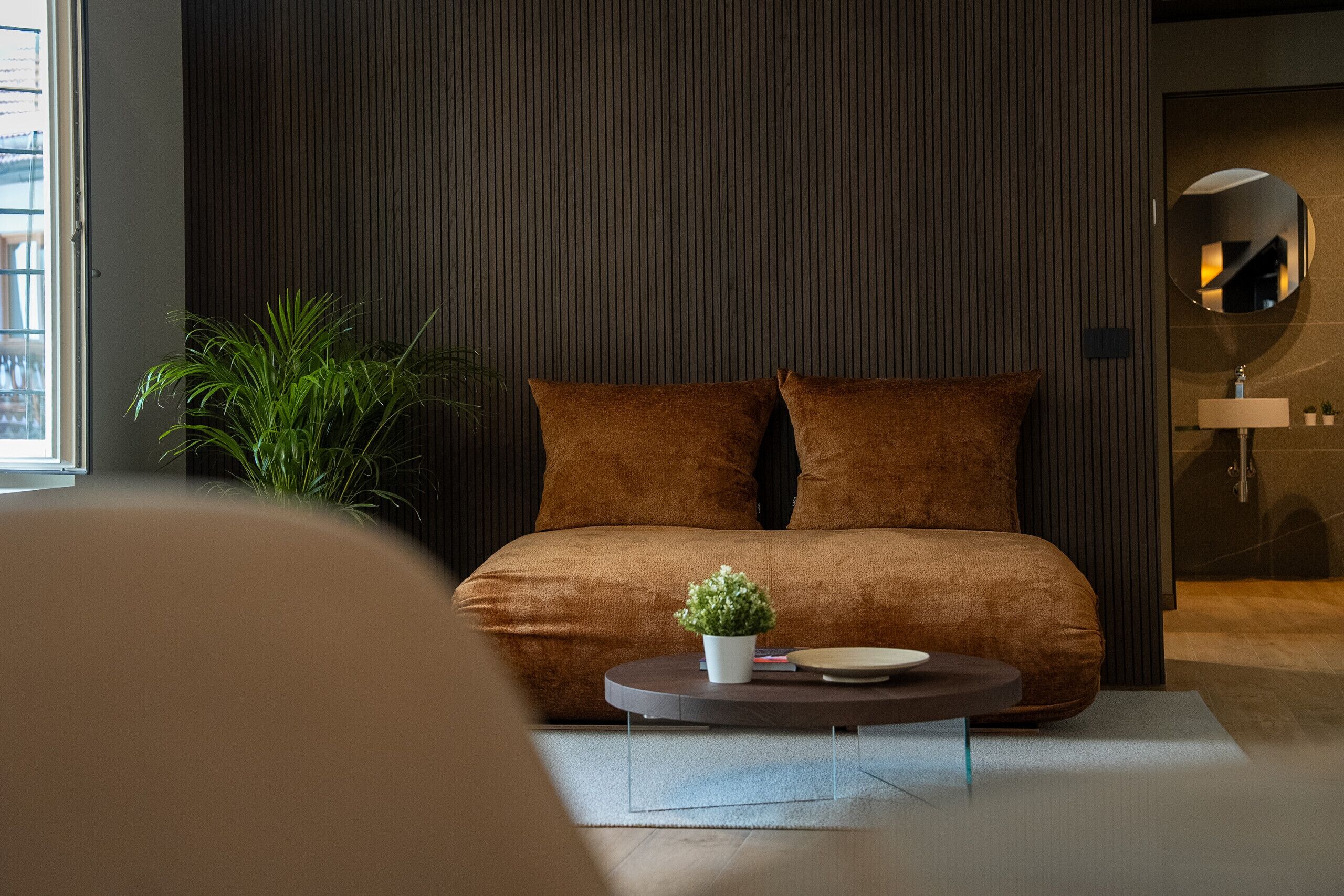 A modern bedroom with a large bed, a small round coffee table, a potted plant, and a mirror on the wall.