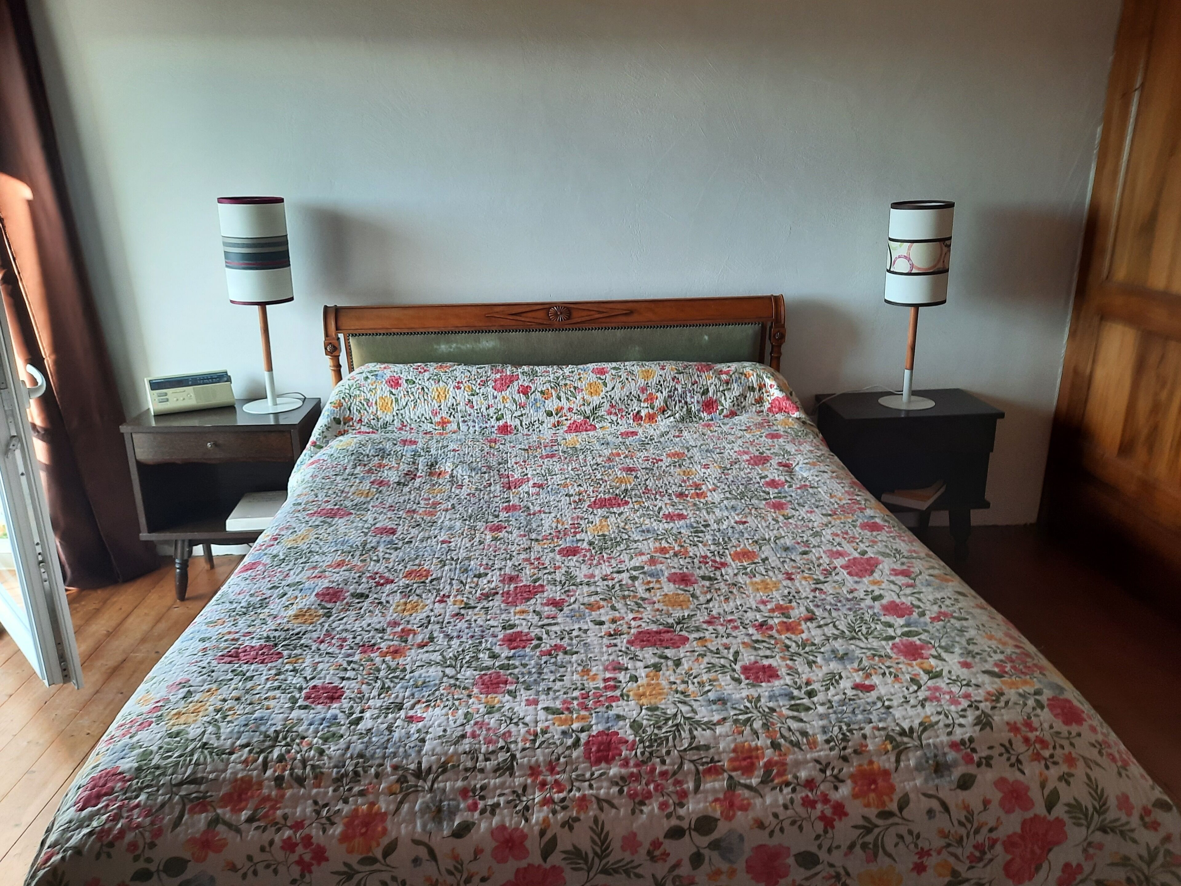 Un dormitorio con colcha estampada, cabecero de madera, dos mesitas de noche con lámparas y una ventana con cortinas.