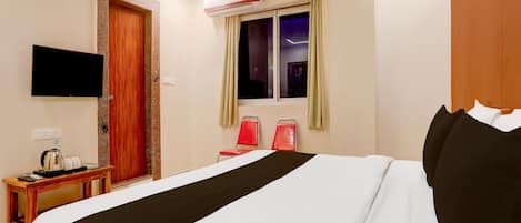 A hotel room with a bed, a TV mounted on the wall, a window with curtains, an air conditioning unit, and a small table with a kettle and cups.