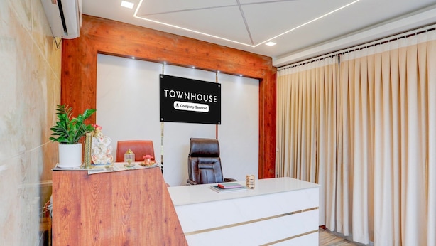 A reception desk with a sign reading 'TOWNHOUSE Company Serviced,' a potted plant, a chair, and a window with curtains. - Townhouse Sri Padmavati Ammavaari Temple (Tirupati)