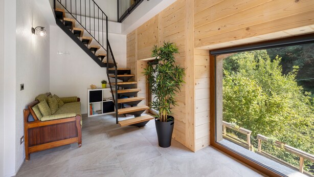 Ein modernes Interieur mit Treppe, Sofa, Topfpflanze und großem Fenster mit Blick auf Bäume.