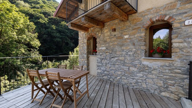 A wooden deck with a table and chairs, stone walls, and a balcony overlooking a forested area. - Faunet Apartment with Mountain View, Wi-Fi and Air Conditioning (Bernezzo)