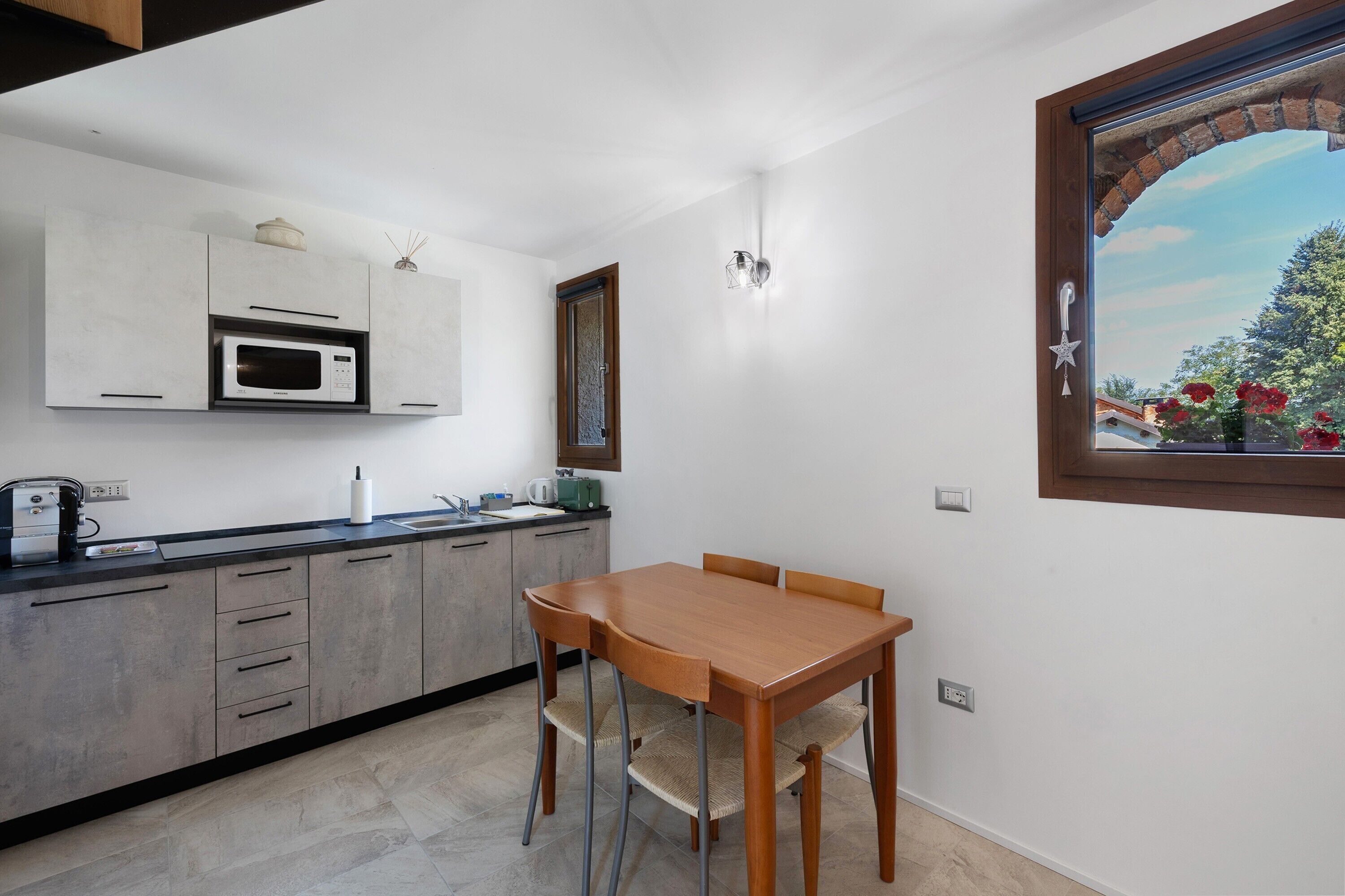 A modern kitchen with a microwave, toaster, and sink. There is a dining table with chairs and a window with a view of flowers.