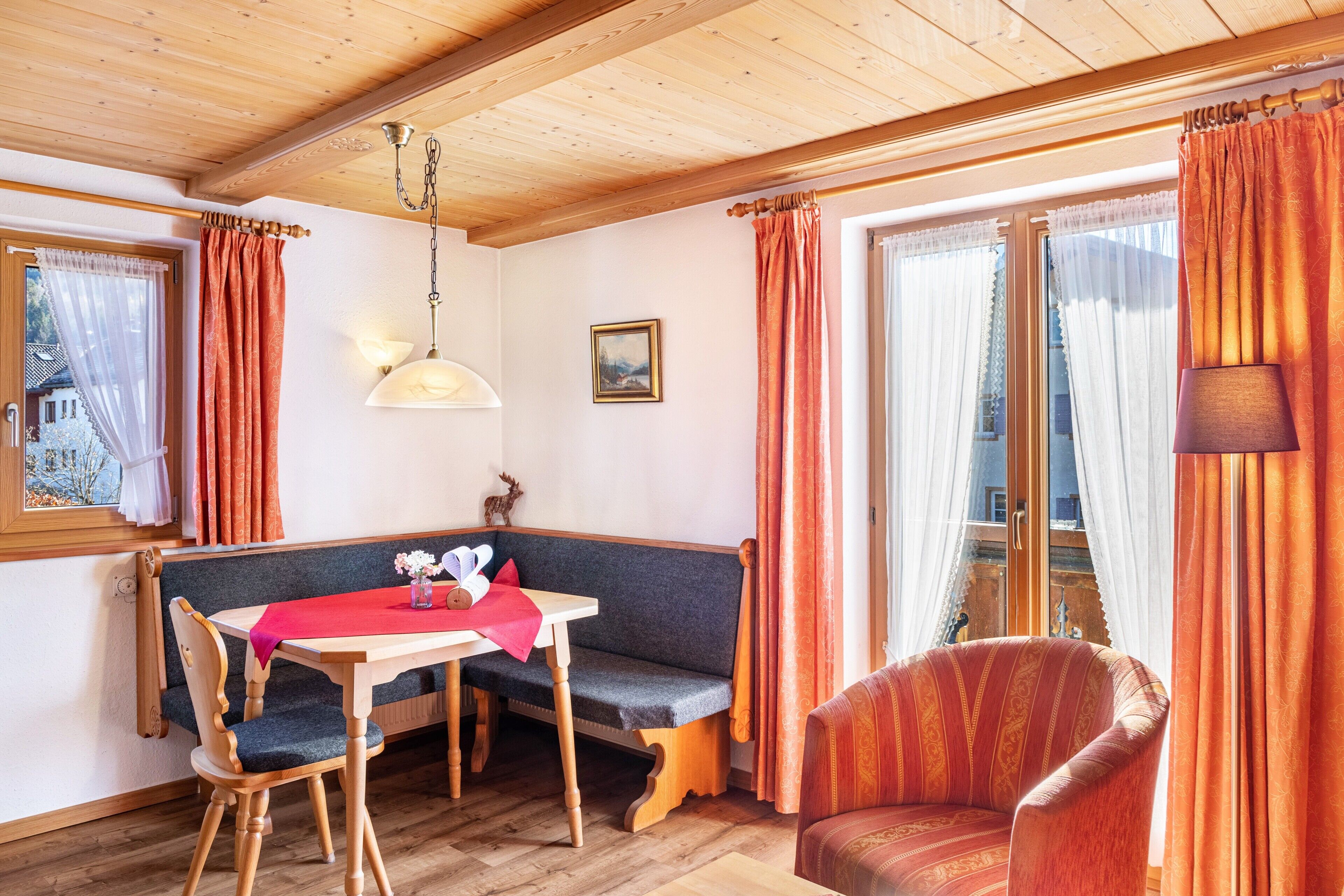 Ein gemütliches Zimmer mit Holzbalkendecke, einer Bank, einem Tisch mit roter Tischdecke und einem gemusterten Sessel.