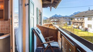 A balcony with a wooden chair, a view of a mountain, and a building with a sloped roof. - Apartment '2' with Mountain View, Balcony and Wi-Fi (Krün)
