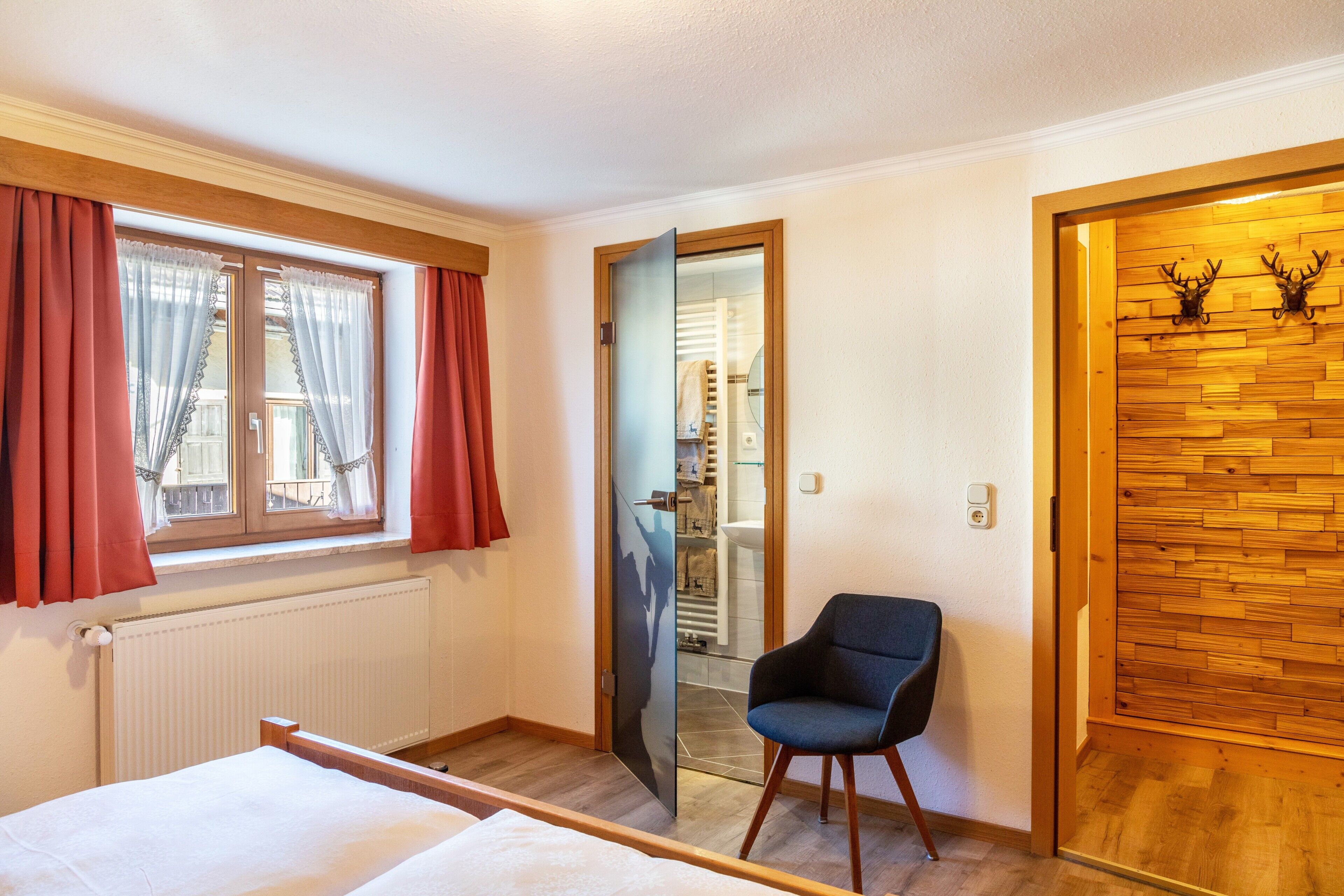 Ein Hotelzimmer mit Bett, Stuhl, einem Fenster mit Vorhängen, einer Holztür mit Geweihen und einem Spiegel.