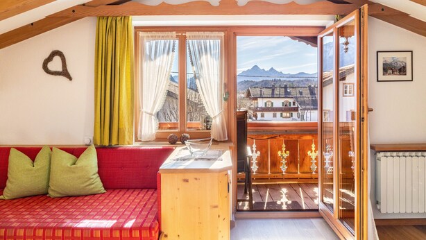 A cozy room with a red sofa, wooden beams, a balcony with a view of mountains, and a wooden cabinet.