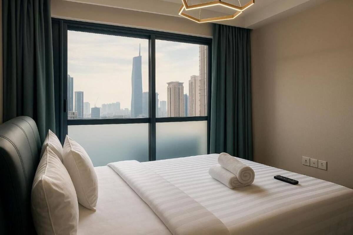 A hotel room with a large window, a bed with white bedding, a remote control, and a view of a cityscape.